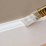 Paintbrush applying white paint to baseboard trim, illustrating how to refresh trim and molding without renovation.