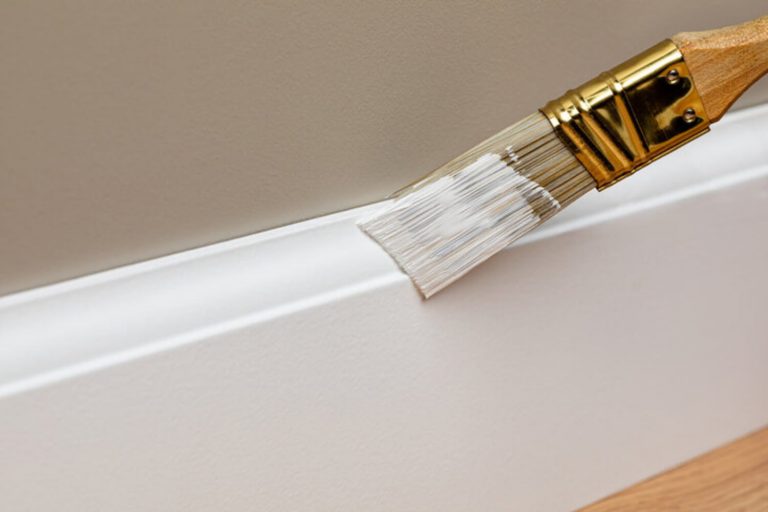 Paintbrush applying white paint to baseboard trim, illustrating how to refresh trim and molding without renovation.