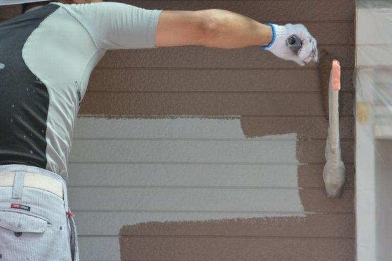 Painter applying brown exterior paint to horizontal siding, illustrating what to expect during exterior house painting in Warwick.
