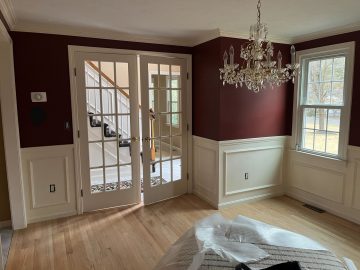 A room highlighted by a striking chandelier against dark walls, framed with white molding, ready for **interior painting services** to refresh. The two windows fill the space with outdoor light, illuminating the potential for **interior painting near me** to transform this area. An empty floor awaits the touch of **interior house painters near me**, making it a canvas for the latest design trends.